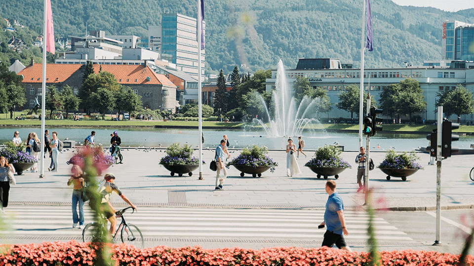 Kuva Bergenin keskustan Festplassenista. Kuvassa on ihmisiä kävelemässä ja pyöräilemässä aukion ja suojatien yli. Paikka on päällystetty kivellä, ja taustalla on vettä. Vedessä on vesilähde, joka luo kuvaan liikettä.